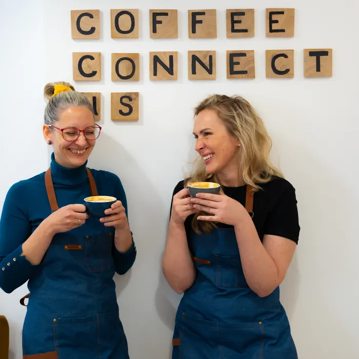 Két barista nevet a „Let coffee connect us” felirat előtt